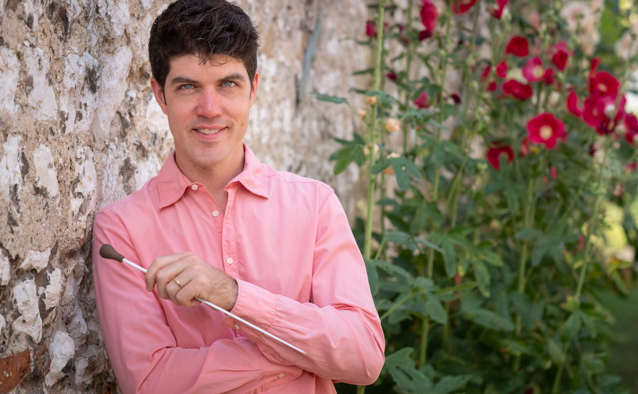 Christopher Quentin McMullen-Laird holding a conductor's baton, leaning against a stone wall with hollyhocks in bloom